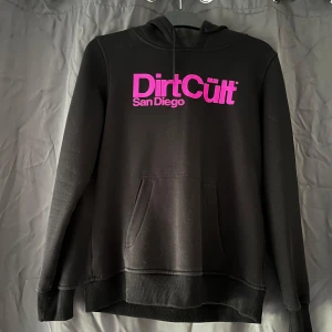 Svart DirtCult hoodie - Svart hoodie från Dirt Cult med stor magficka och huva. Framsidan har ett stort rosa tryck med texten 'Dirt Cult San Diego'. Tröjan har långa ärmar och en klassisk passform, perfekt för chill dagar. Materialet känns mjukt och skönt mot huden. Aldrig använd, bara provad. Den är väldigt liten i sin storlek och hade kunnat klassas som en S/M. Har inte kvar orginalförpackning. 