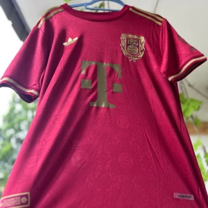 FC Bayern München 125 år Adidas tröja - Exklusiv FC Bayern München 125-årsjubileumströja från Adidas. Djup röd färg med guldiga detaljer, broderat klubbmärke och Adidas-logga. Klassisk passform med korta ärmar och subtila mönster i tyget. Tillverkad i lätt och ventilerande material (AEROREADY). Alla storlekar tillgängliga, tryck löses gratis vid special beställning och alla tröjor i världen finns, allt från irakiska landslaget till Real Madrid !