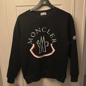 Svart Moncler sweatshirt  - Svart sweatshirt från Moncler med stor vit och röd logga framtill och broderad patch på ärmen. Klassisk rund halsringning och ribbade muddar. Perfekt för dig som gillar streetstyle och vill sticka ut med ett exklusivt märke.