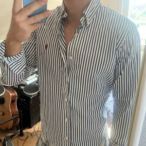 Randig skjorta från Ralph Lauren - Säljer en klassisk vit skjorta med marinblå vertikala ränder från Ralph Lauren. Skjortan har lång ärm, knappar hela vägen och ikoniska röda broderade loggan på bröstet. Snygg passform och tillverkad i mjuk bomull. Perfekt till jeans eller chinos.
