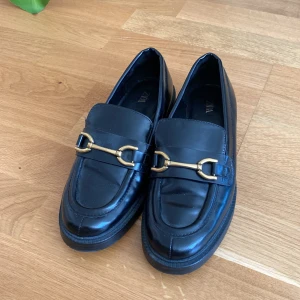 Svarta loafers med gulddetalj från Zara - Stilrena svarta loafers från Zara, köpta på second hand, med chunky sula och snygg guldfärgad metallspänne framtill. Skorna är i glansigt skinnliknande material och har en klassisk, rund tå. Perfekta för en dressad look med edge. Skorna är något slitna och använda av mig ca 2 ggr. 
