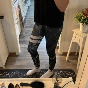 Svart gula Stronger leggings med mönster - Säljer ett par leggings från Stronger med coolt marmorerat mönster i svart grått, vitt och gult. Hög midja med logotyp i midjebandet och vita ränder på ena benet. Sitter tight och är perfekta för träning.