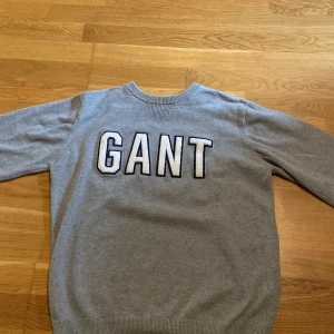 Grå GANT tröja med stor logga - Snygg grå tröja från GANT i bomull med stor vit och blå logga framtill. Klassisk rund halsringning och ribbade muddar vid ärmslut och nederkant. Perfekt för dig som gillar stilren och sportig look.