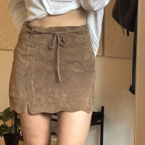 Brun mockakjol med snörning - Supersöt brun kortkjol i mocka med snörning framtill och vågig nederkant. Kjolen har en snygg, figurnära passform och coola sömdetaljer längs sidorna. Perfekt för dig som gillar en boho eller vintage vibe.