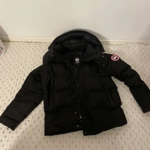 Svart dunjacka från Canada Goose - Säljer en svart dunjacka från Canada Goose med huva och klassisk logga på ärmen. Jackan har dragkedja och knappar framtill, samt flera praktiska fickor. Finns även päls och box. Lite sliten men det syns inte. Pris kan diskuteras. 