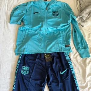 Barca Nike träningset - Snygga marinblå träningsbyxor med FC Barcelona-logga och turkosa detaljer längs benen. Byxorna har elastisk midja med snörning och är tillverkade i ett lätt och ventilerande polyestermaterial. Perfekta för fotbollsträning eller chill. Tröjan är torkosblå med en dragkedja hela vägen ner och upp. Ingeting är använt därför är de bästa skick på allting. Man kan också köpa bara tröjan eller bara byxorna men hör av er vid frågor! Pris kan diskuteras vid snabb affär. 