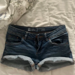 Lågmidjade jeansshorts i blå denim - Snygga lågmidjade jeansshorts från C Denim i klassisk blå denim. Modellen har upprullade benslut, fem fickor och dragkedja med knapp. Perfekta för varma dagar och en avslappnad stil.