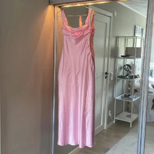 Ljusrosa långklänning i satin - Supersnygg ljusrosa långklänning i glansigt satinmaterial med breda axelband och draperad urringning. Klänningen har en figurnära passform och axelbanden kan bäras både på axeln och offshoulder för en cool look.