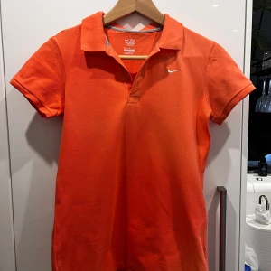 Orange pikétröja från Nike - Snygg orange pikétröja från Nike med klassisk krage och korta ärmar. Tröjan har en liten vit Nike-logga broderad på bröstet och är gjord i bomull för en skön känsla. Perfekt för dig som gillar sportig och stilren look.