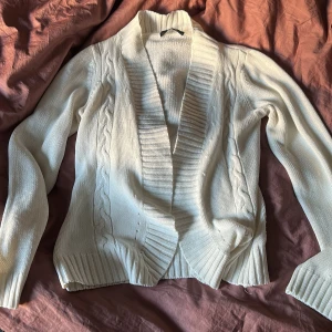 Vit cardigan från Vero Moda - Mysig vit cardigan jacka från Vero Moda med kabelstickade detaljer och bred ribbad kant. Jackan är långärmad, öppen framtill och har en klassisk, enkel look som passar perfekt till jeans eller klänning. Perfekt för lager-på-lager under kyliga dagar.
