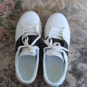 Valentino Garavani Open sneakers vit/svart - !PRUTA GÅR! Använda 3 gånger. Snygga Valentino Garavani Open sneakers i vitt skinn med bred svart läderdetalj över vristen. Klassisk rund tå, vita snören och coola nitar längs sulan. Perfekt för dig som vill ha en stilren men ändå lyxig look.