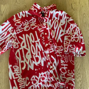 Röd oversized skjorta från FSBN - Säljer en röd oversized skjorta från FSBN med vit graffiti-inspirerad text över hela plagget. Skjortan har korta ärmar, knappar framtill och en riktigt streetig vibe. Perfekt för dig som vill sticka ut med en unik look.