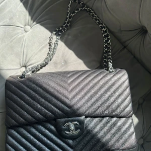 Svart axelväska i skinn - Säljer en klassisk svart axelväska i kornigt skinn med silverfärgad kedja och ikoniskt CC-spänne. Väskan har quiltad design i chevronmönster och röd insida med broderad logga. Perfekt storlek för det viktigaste och riktigt snygg detalj till din outfit.