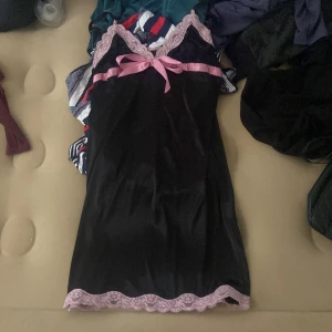 Svart kort klänning med rosa spets - Supersöt svart kort klänning från Secret med tunna axelband, rosa spetsdetaljer och en stor rosa rosett framtill. Klänningen har en glansig satinliknande yta och är perfekt för dig som gillar en feminin och lekfull stil.