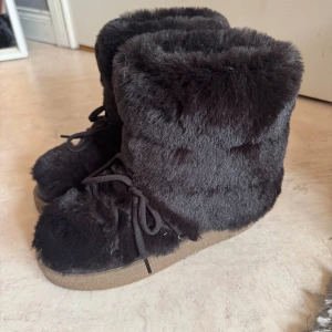 Svarta fluffiga boots med snörning - Mysiga svarta boots med fluffig pälsimitation och snörning framtill. Skorna har en rund tå och platt sula i gummi, perfekta för kalla dagar. De är höga och täcker ankeln för extra värme och stil.