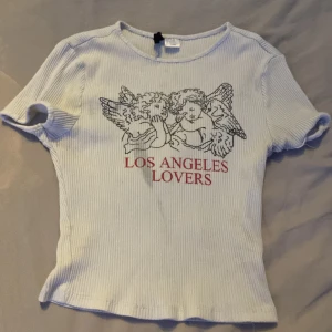 Vit ribbad topp med ängeltryck - Vit ribbad topp med korta ärmar och tryck av två änglar samt texten 'Los Angeles Lovers' i rött framtill. T-shirten har en tight passform och är tillverkad i mjukt bomullsmaterial. Perfekt för dig som gillar coola prints och streetstyle. OBS! den har en rätt så stor fläck under bröstet.