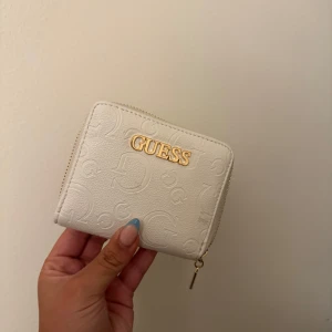 Vit plånbok från Guess med guldiga detaljer - Supersnygg vit plånbok från Guess med präglade loggor och guldfärgad dragkedja. Insidan har flera kortfack och myntficka. Perfekt storlek för att ha med sig överallt och ger en clean och lyxig känsla. Har använt den en gång bara! 