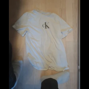 Vit t-shirt från Calvin Klein - Vit t-shirt från Calvin Klein med klassisk svart CK-logga på bröstet. Enkel och clean design med rund hals och korta ärmar. Perfekt för dig som gillar stilrena plagg och vill ha något som funkar till det mesta. Passar S