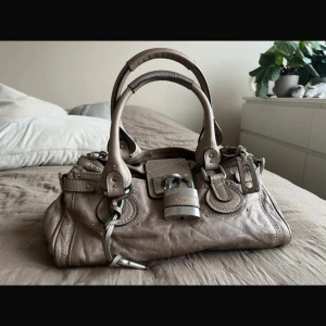 Chloé Paddington handväska i beige skinn - Säljer en ikonisk Chloé Paddington handväska i beige skinn med silverfärgade metalldetaljer och det klassiska hänglåset med tillhörande nyckel. Väskan har dubbla handtag, dekorativa sömmar och dragkedja upptill. Perfekt för dig som gillar statement-accessoarer.