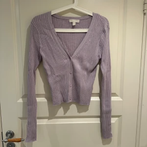 Ljuslila ribbad kofta med v-ringning - Superfin ljuslila kofta från H&M med ribbad struktur och v-ringad hals. Modellen är croppad och har långa ärmar samt knappar framtill. Perfekt att styla med jeans eller kjol för en trendig look.