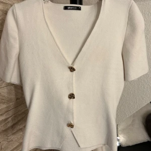 Vit v-ringad topp från Gina Tricot - Vit v-ringad topp från Gina Tricot med korta ärmar och tre unika knappar framtill. Toppens material känns mjukt och har en lätt stickad känsla. 