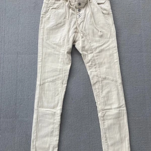 Beige Please byxor med snörning - Snygga beige byxor från Please med smal passform, hög midja och cool snörning i midjan. Byxorna har dekorativa sömmar, bakfickor och ett litet hjärtformat metallmärke bak. Perfekta för en avslappnad men trendig look.