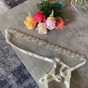 Glittrigt silverfärgat midjebälte - Supersnyggt midjebälte med glittriga stenar och pärlor i silverton. Bältet har ett brett band med broderade detaljer och knyts med ett ljusvitt satinband. Perfekt accessoar för att lyfta din outfit med extra bling.