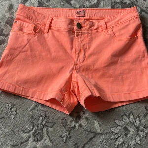 Korta korallrosa shorts från ONLY - Snygga korallrosa shorts från ONLY med klassisk femficksdesign och knappstängning. Modellen är kort och har bälteshällor, perfekt för varma dagar. Materialet är mjukt och skönt bomullstyg som andas. Enkla att matcha med allt i garderoben.