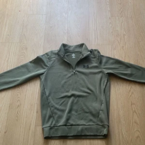 Olivgrön fleece-tröja från Under Armour - Snygg olivgrön långärmad fleece-tröja från Under Armour med svart logga på bröstet. Tröjan har half zip och hög krage, perfekt för kyliga dagar. Mjuk insida i fleece och normal passform. Passar både till träning och chill.