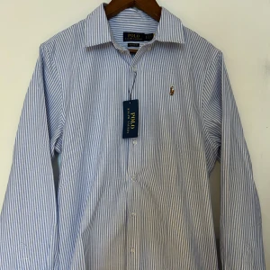 Randig skjorta från Polo Ralph Lauren - Klassisk skjorta från Polo Ralph Lauren i blåvitt randigt mönster. Skjortan har lång ärm, knappar framtill och broderad logga på bröstet. Tillverkad i bomull och har en klassisk passform. Perfekt för dig som gillar stilren och tidlös design. Pris kan diskuteras vid snabb affär 