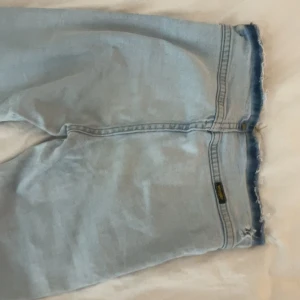 Bootcutjeans  - Ljusblå bootcutjeans från Wrangler med råa, fransiga kanter och slitna detaljer. Klassisk femficksmodell i denim med avslappnad passform. Perfekta för en chill sommarlook och funkar till allt från sneakers till sandaler.