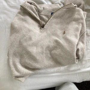 Beige half zip tröja Polo Ralph Lauren - Snygg beige långärmad tröja från Polo Ralph Lauren med klassisk broderad logga på bröstet. Tröjan har en half zip-dragkedja vid kragen och är tillverkad i mjuk bomull. Perfekt för en chill och stilren look.