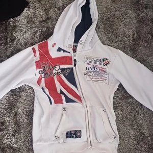 Vit hoodie från Geo Norway med tryck - Säljer en vit hoodie från Geo Norway med Union Jack-tryck och broderade detaljer på framsidan. Tröjan har huva, dragkedja och fickor framtill. Perfekt för dig som gillar sportig och streetinspirerad stil.