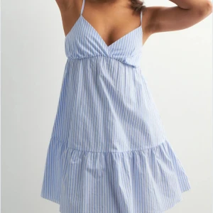 Blåvit randig kort sommarklänning - Supersöt kort klänning med tunna axelband och v-ringning. Klänningen är blå- och vitrandig med volang nertill.