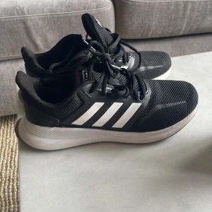 Svarta Adidas löparskor  - Snygga svarta Adidas sneakers med klassiska vita ränder på sidorna och vit sula. Ovandelen är i mesh för bra andningsförmåga och skorna har snörning. Perfekta för dig som gillar sportig och clean stil.