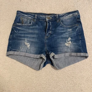 Blå jeansshorts med slitningar - Snygga blå jeansshorts med uppvikta benslut och slitna detaljer framtill och baktill. Klassisk femficksmodell med dragkedja och knapp. Perfekta för en chill sommardag och ger en avslappnad vibe.