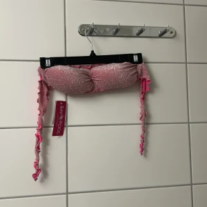 Glittrig rosa bikiniöverdel Baypool - Superfin glittrig rosa bikiniöverdel från Baypool med bandeau-modell och långa knytband med volangkant. Perfekt för att sticka ut på stranden eller vid poolen. Snygg och trendig design med metallic-effekt och rynk i mitten.