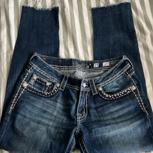 Mörkblå jeansbyxor från Miss Me med dekorativa detaljer - Säljer ett par mörkblå jeans från Miss Me med snygga slitningar och dekorativa sömmar samt nitar på fickorna. Byxorna har raka ben med råa, avklippta benslut och klassisk femficksdesign. Perfekta för dig som gillar detaljer och unik stil.