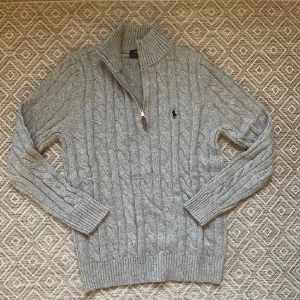 Grå kabelstickad tröja med dragkedja från Ralph Lauren - Säljer en grå kabelstickad tröja från Ralph Lauren med halv dragkedja och hög krage. Tröjan har långa ärmar och klassisk broderad logga på bröstet. Använd cirka 2 månader. Skriv vid funderingar!