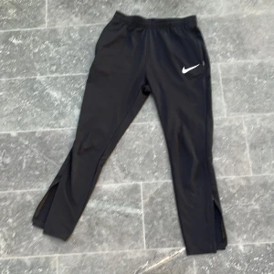 Svarta träningsbyxor från Nike Dri-Fit - Svarta träningsbyxor från Nike med Dri-Fit-teknologi. Byxorna har luftig passform blandat med tajt, dragkedjor vid bensluten och snörning i midjan. Perfekta för träning och sportiga aktiviteter. Klassisk Nike-logga på låret.
