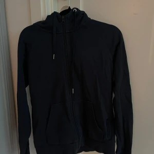 Svart hoodiejacka från Peak Performance - Svart hoodiejacka från Peak Performance med dragkedja framtill och två fickor. Jackan har huva med dragsko och är tillverkad i mjukt material, perfekt för vardagsbruk eller träning.