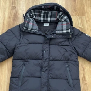 Svart dunväst från Burberry med huva - Säljer en svart dunväst från Burberry med avtagbara ärmar och huva. Västen har två dragkedjefickor och ett klassiskt rutigt foder i grått, vitt och rött. Perfekt för lager-på-lager och snygg streetstil. Har bara använd den få gånger. Storleken på den är M. Säljer den då den inte passar mig längre. Priset på jackan kommer vara 2599kr, men kan diskuteras vid snabb köp 