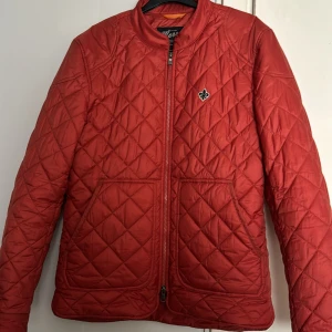 Röd quiltad jacka från Morris - Säljer en röd quiltad jacka från Morris med dragkedja framtill och två stora fickor. Jackan har ett diskret emblem på bröstet och en stilren, klassisk look. Perfekt för dig som vill ha en lätt och snygg jacka till vardags. Nypris på dessa jackor är 3000kr. Har 2 små fläckar på ryggen man ser det på bilden.
