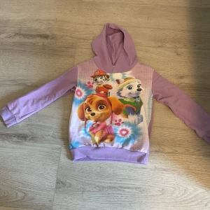 Lila Paw Patrol hoodie med tryck - Säljer en söt lila hoodie med Paw Patrol-tryck framtill. Tröjan har huva och långa ärmar, samt färgglada motiv av karaktärerna Skye, Everest och Marshall. Perfekt för dig som gillar mjuka och bekväma tröjor.