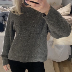 Grå stickad tröja med pärlor - Mysig grå stickad tröja med små pärlor framtill. Tröjan har lång ärm och rund halsringning, perfekt för kyligare dagar. Enkel och stilren design som passar till mycket.