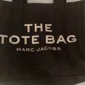 Svart tote bag från Marc Jacobs - Säljer en svart tote bag från Marc Jacobs med tydlig vit text på framsidan. Väskan har både handtag och en avtagbar axelrem, perfekt för att bära på olika sätt. Klassisk och rymlig modell med patchdetalj på baksidan.