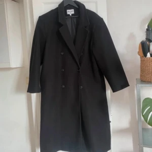 Svart kappa från NA-KD - Säljer en svart, lång kappa från NA-KD i oversized modell. Kappan har dubbelknäppning framtill med svarta knappar och klassisk krage. Perfekt för dig som gillar stilrena och tidlösa plagg.