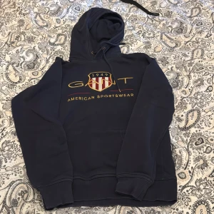 Mörkblå hoodie från GANT - Säljer en mörkblå hoodie från GANT med broderad logga och texten 'American Sportswear' på bröstet. Tröjan har huva med dragsko och ribbade muddar vid ärmslut och nederkant. Perfekt för en avslappnad stil.