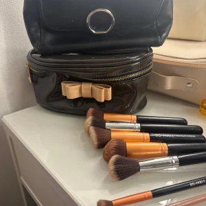 Set med sminkborstar och sminkväskor - Pris kan diskuteras! Flera sminkborstar med svarta och orange handtag, täta och mjuka borst. Två sminkväskor i svart, en med guldfärgad rosett och dragkedja, samt en stilren svart clutch med silverdetalj. Perfekt för att organisera och applicera smink. 