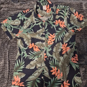 Blommig kortärmad skjorta från Lindex - Säljer en färgglad kortärmad skjorta från Lindex med tropiskt blommönster i grönt, orange och beige på mörk botten. Skjortan har klassisk krage och knäppning framtill. Perfekt för dig som gillar en somrig och avslappnad stil!